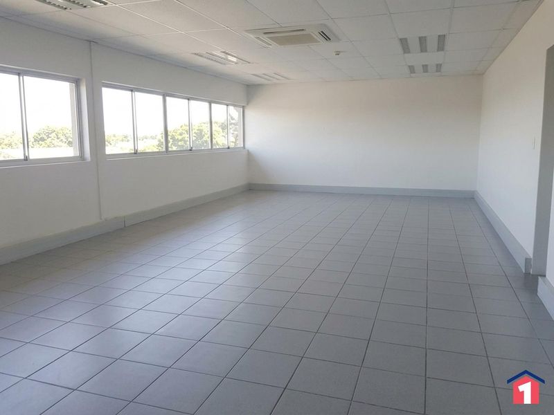 A spacious, empty office area featuring tiled flooring, a suspended ceiling with recessed lighting, and large windows providing ample natural light.