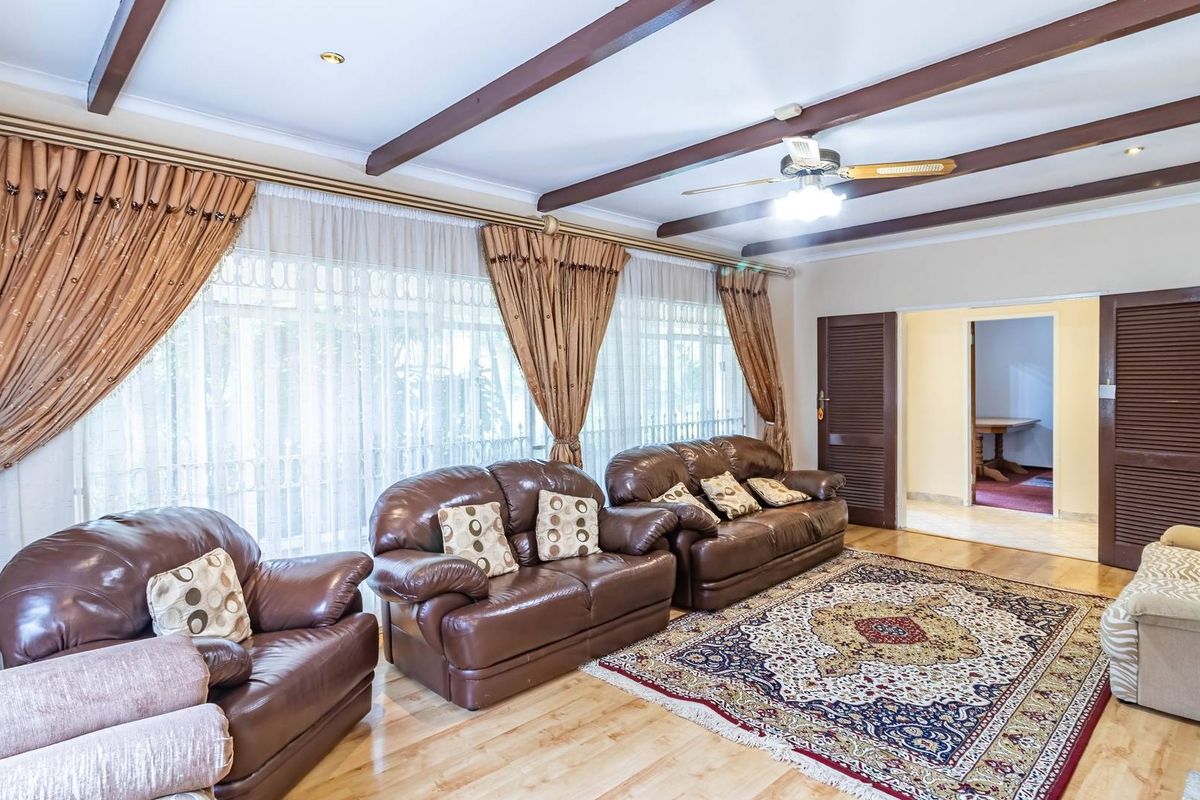Spacious living room featuring exposed timber ceiling beams, light wood flooring, and large windows with floor-to-ceiling curtains, leading to an adjacent room.
