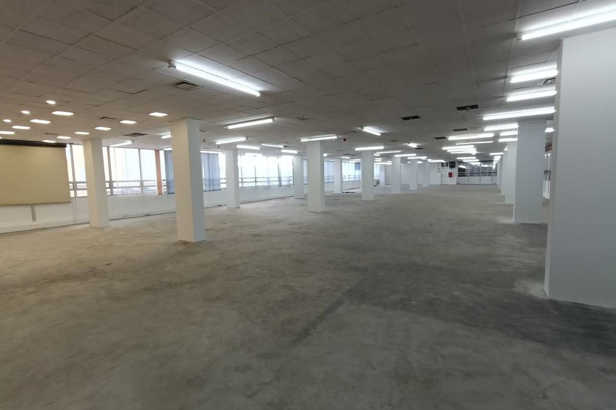 A large, open-plan office space featuring concrete flooring, structural columns, and a suspended ceiling with integrated fluorescent lighting.