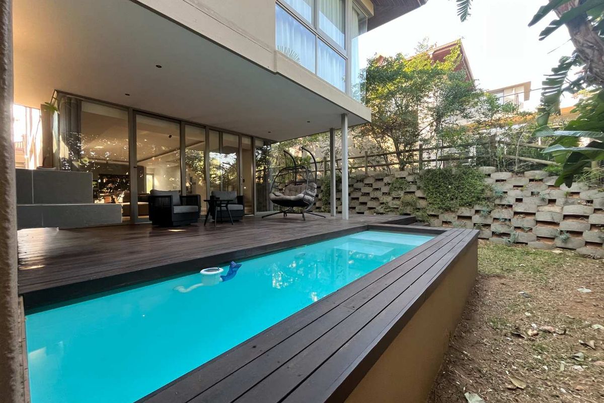 A modern outdoor living space featuring a swimming pool with timber decking, a covered patio area, and a landscaped retaining wall.