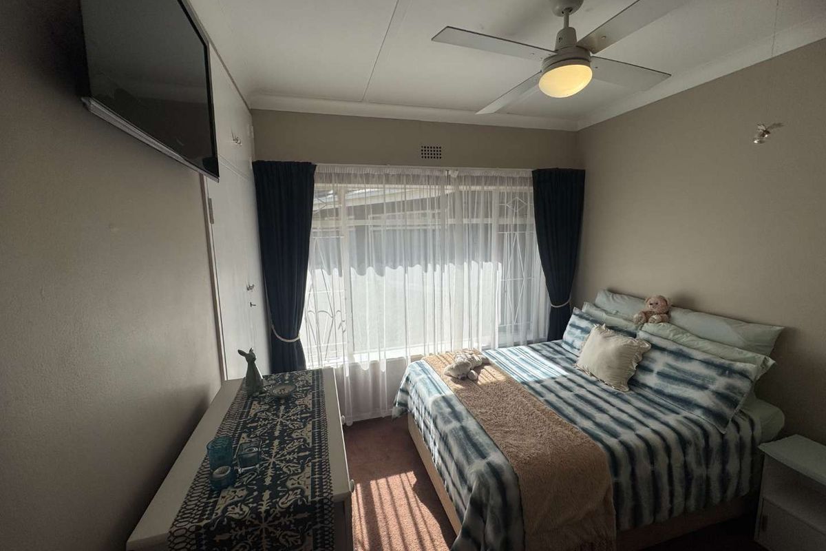 A cozy bedroom featuring a ceiling fan, large window with security bars, and neutral wall finishes.