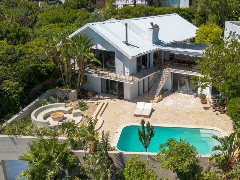 Aerial view of a multi-level residence featuring a swimming pool, stone patio, built-in fire pit, and an external staircase leading to an upper balcony.