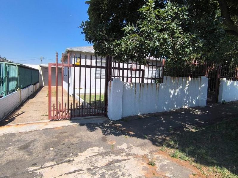 Front exterior view of the property featuring a paved driveway, metal security gate, and perimeter wall with visible signs of weathering and rust.