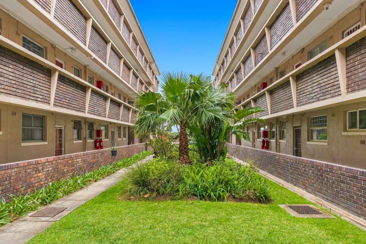 A view of the central courtyard garden between two apartment blocks, featuring brick facades, manicured lawn, and established greenery.