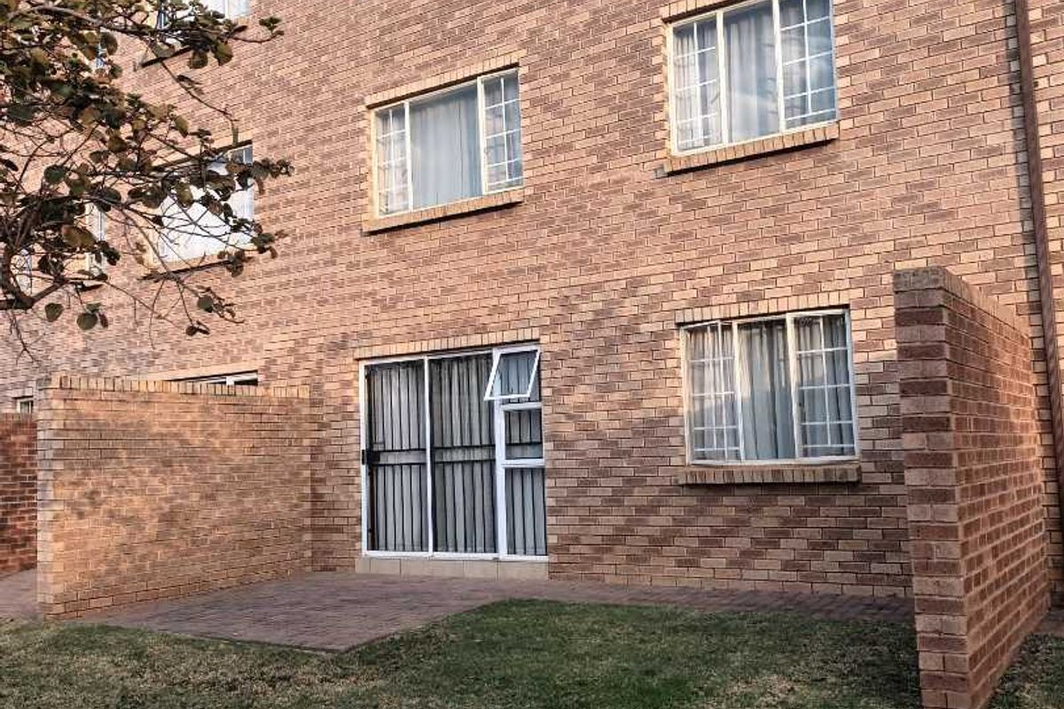 Rear view of a multi-story brick apartment building featuring a ground-floor patio area with a small private lawn and security-fitted sliding doors.