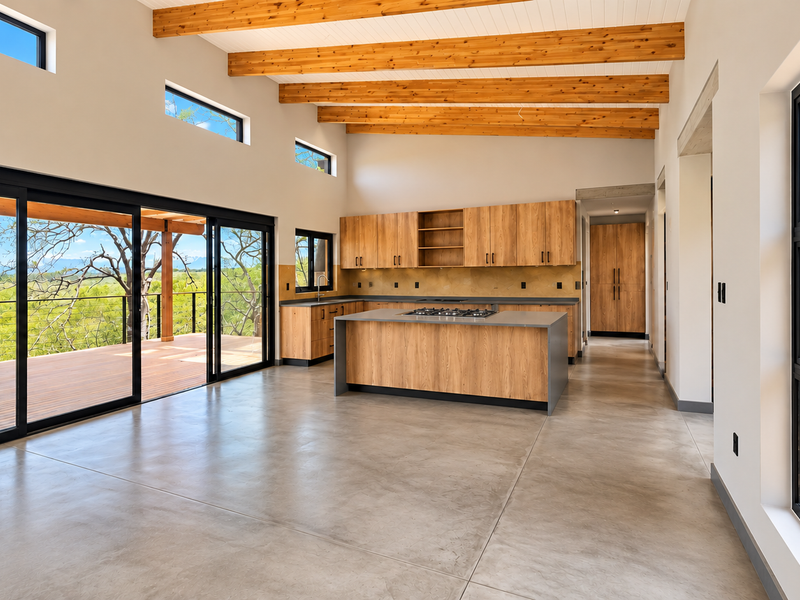 Spacious open-plan kitchen and living area featuring polished concrete floors, exposed timber beams, modern wooden cabinetry, and large sliding doors opening to a deck.