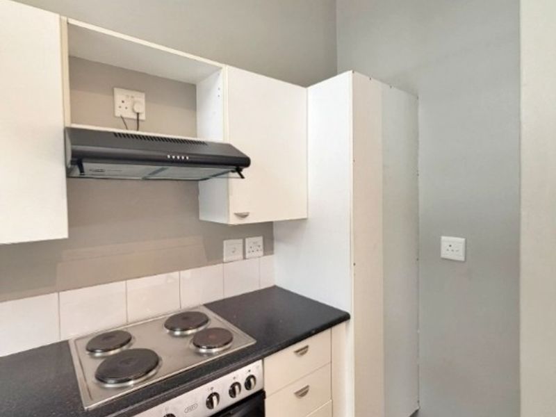 A compact kitchen featuring white built-in cabinetry, dark laminate countertops, an integrated oven, electric hob, and extractor fan with neutral tiled flooring.