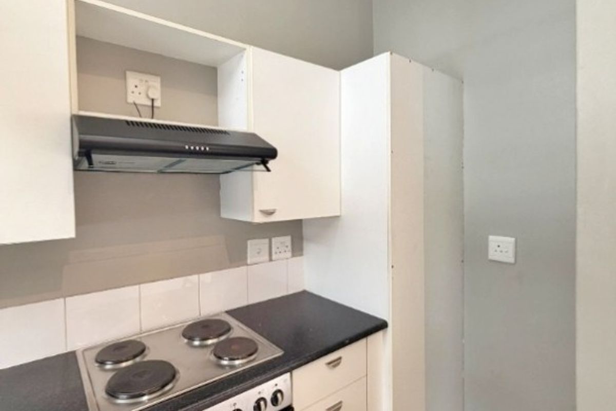 A compact kitchen featuring white built-in cabinetry, dark laminate countertops, an integrated oven, electric hob, and extractor fan with neutral tiled flooring.