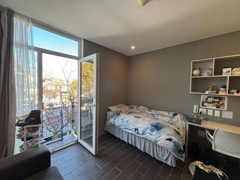A modern bedroom featuring tiled flooring, built-in desk and shelving, and direct access to a private balcony through a glass door.
