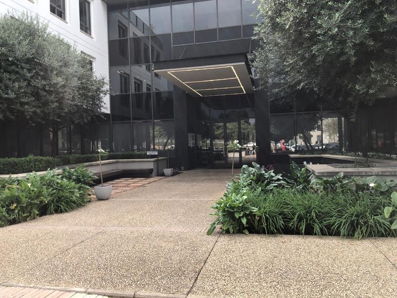 Modern office building entrance featuring a glass facade, sleek canopy with integrated lighting, and well-maintained landscaped garden beds along the paved walkway.
