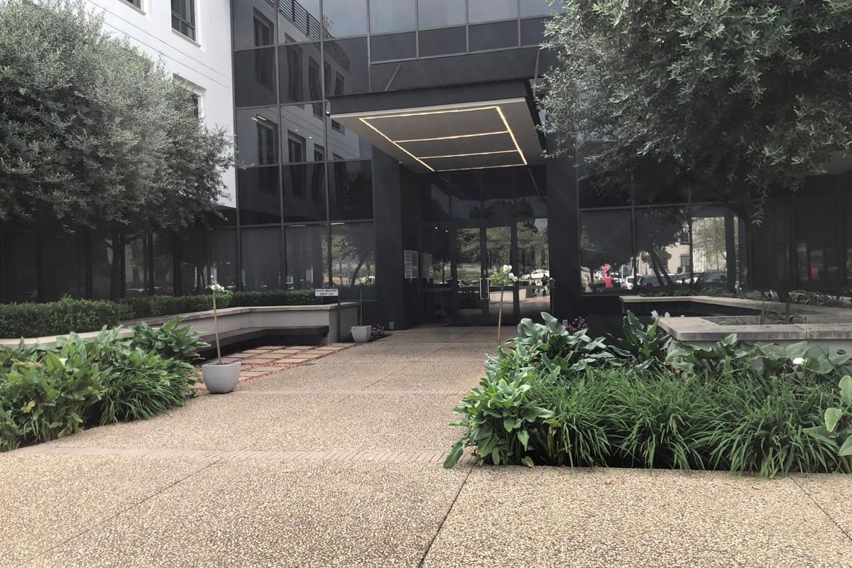 Modern office building entrance featuring a glass facade, sleek canopy with integrated lighting, and well-maintained landscaped garden beds along the paved walkway.