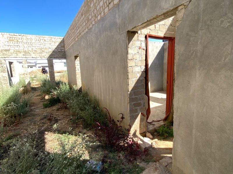 An unfinished residential structure showing exposed concrete block walls, cement plastering, and a metal door frame in a construction site setting.