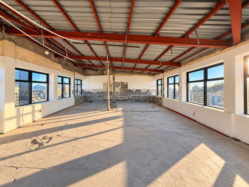 A large, open-plan commercial space in shell condition, featuring exposed steel roof trusses, concrete floors, and expansive windows with city views.