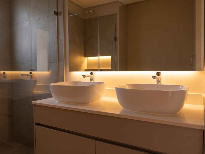 Modern bathroom featuring a double vanity with vessel sinks, a large backlit mirror, and a glass-enclosed shower with contemporary tile finishes.