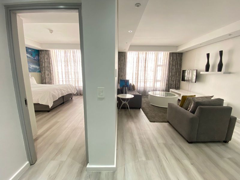 An open-plan living area and bedroom featuring modern laminate flooring, recessed lighting, and large windows with floor-to-ceiling curtains.