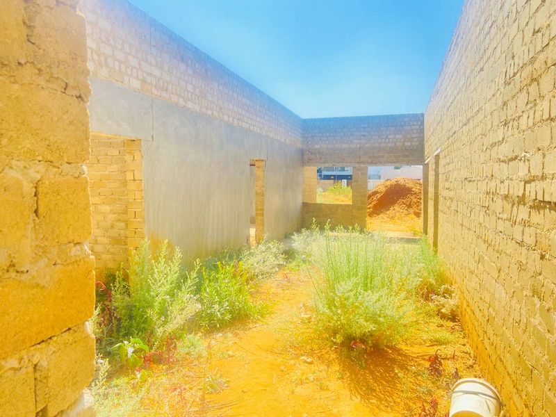 An unfinished construction site showing exposed brick walls and a dirt floor with overgrown vegetation, indicating a project in the early stages of development.