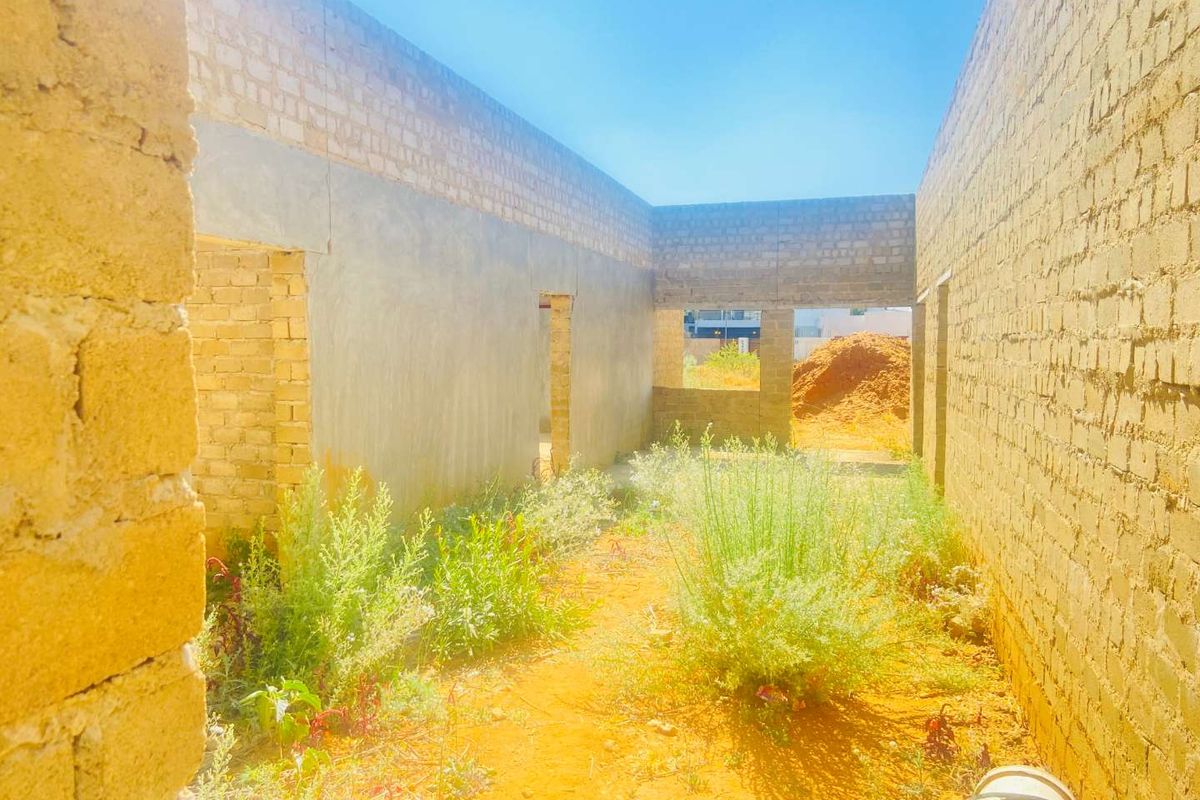 An unfinished construction site showing exposed brick walls and a dirt floor with overgrown vegetation, indicating a project in the early stages of development.