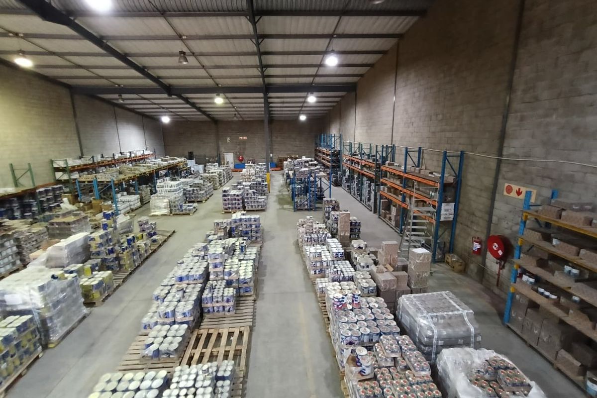 Spacious industrial warehouse featuring high-bay lighting, exposed steel roof trusses, concrete flooring, and extensive built-in steel racking systems for storage.
