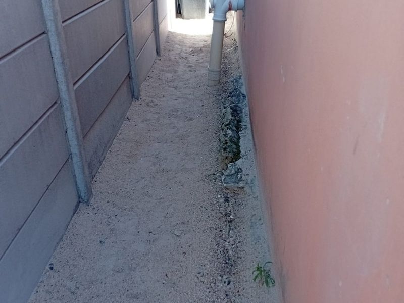 A narrow side passage between a precast concrete boundary wall and the main building structure, featuring exposed foundation work and sandy ground.