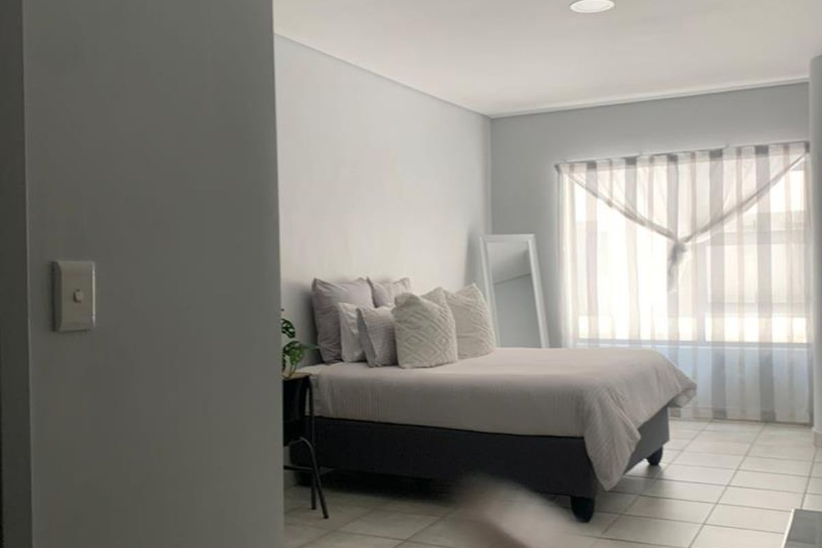 A bedroom featuring tiled flooring, neutral wall finishes, and a large window with sheer curtains, providing a bright and simple living space.