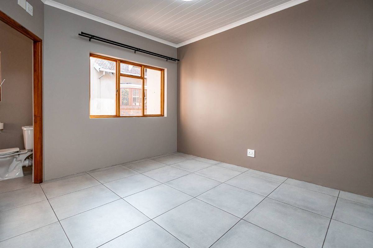 A clean, empty bedroom featuring neutral painted walls, tiled flooring, and a timber-framed window, with an adjacent bathroom visible.