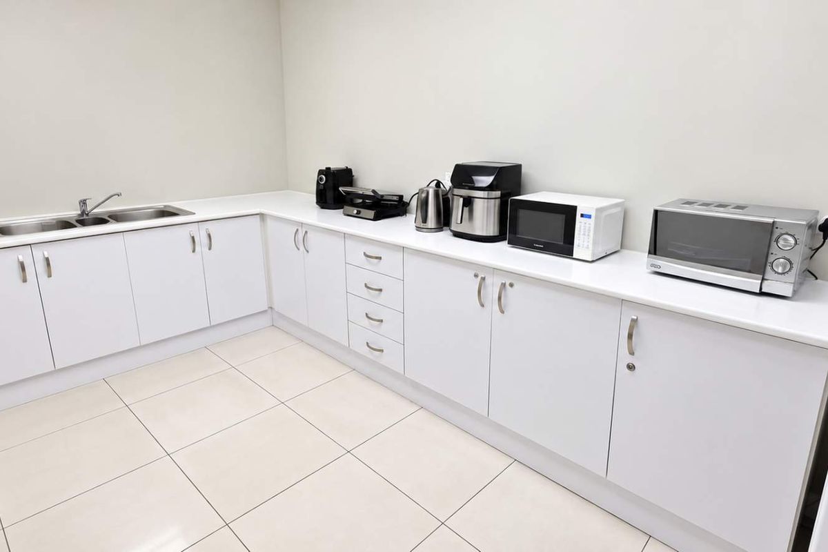 A clean and functional office kitchenette featuring white cabinetry, laminate countertops, a double sink, and tiled flooring.