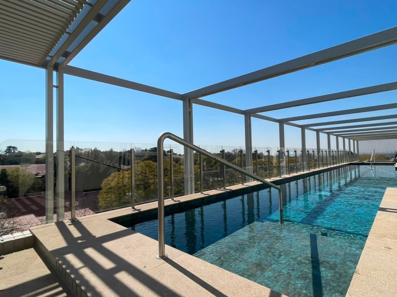 A modern rooftop swimming pool featuring a sleek metal pergola, glass balustrades, and panoramic views of the surrounding area.