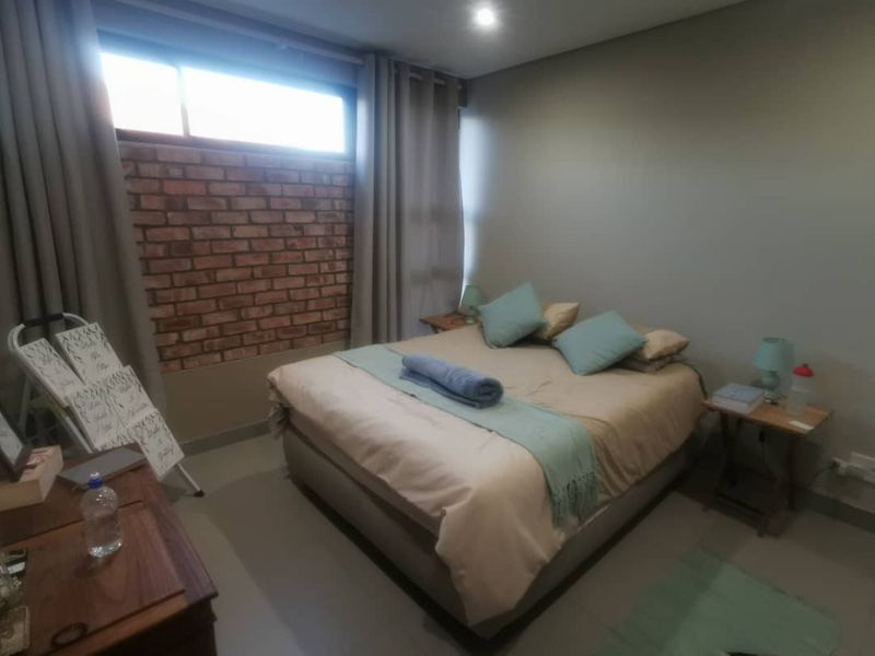 A comfortable bedroom featuring an exposed brick accent wall, neutral tiled flooring, and recessed ceiling lighting.