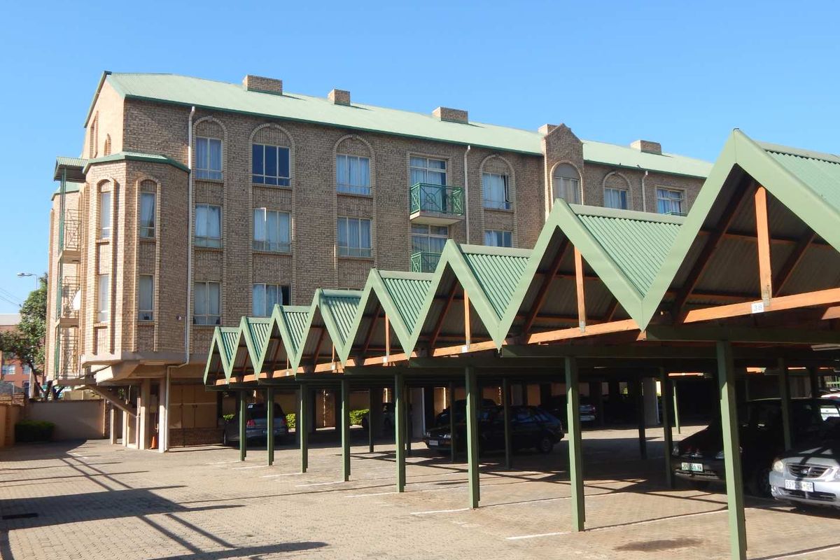 Exterior view of a multi-story brick apartment building featuring a large covered parking area with a distinctive green corrugated roof structure.
