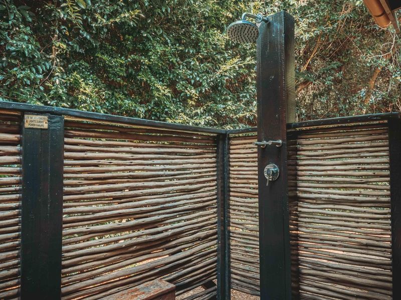 An outdoor shower area featuring a fixed shower head and mixer, enclosed by a rustic timber privacy screen set against a natural garden backdrop.