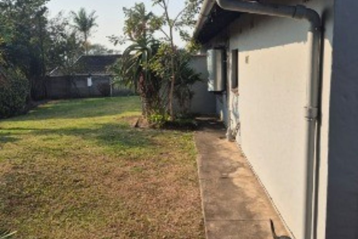 A view of the rear exterior of the house featuring a concrete pathway, guttering system, and an external geyser unit alongside a grassed garden area.