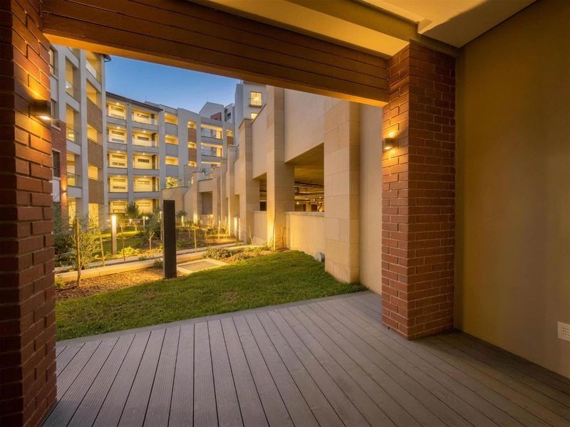 A modern covered patio featuring composite decking and brick pillars, overlooking a well-maintained landscaped garden area within the apartment complex.