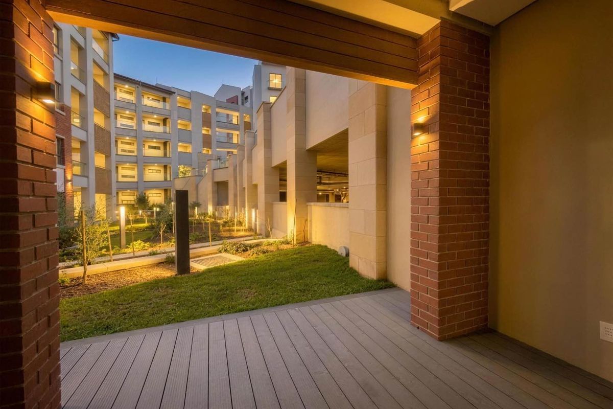 A modern covered patio featuring composite decking and brick pillars, overlooking a well-maintained landscaped garden area within the apartment complex.