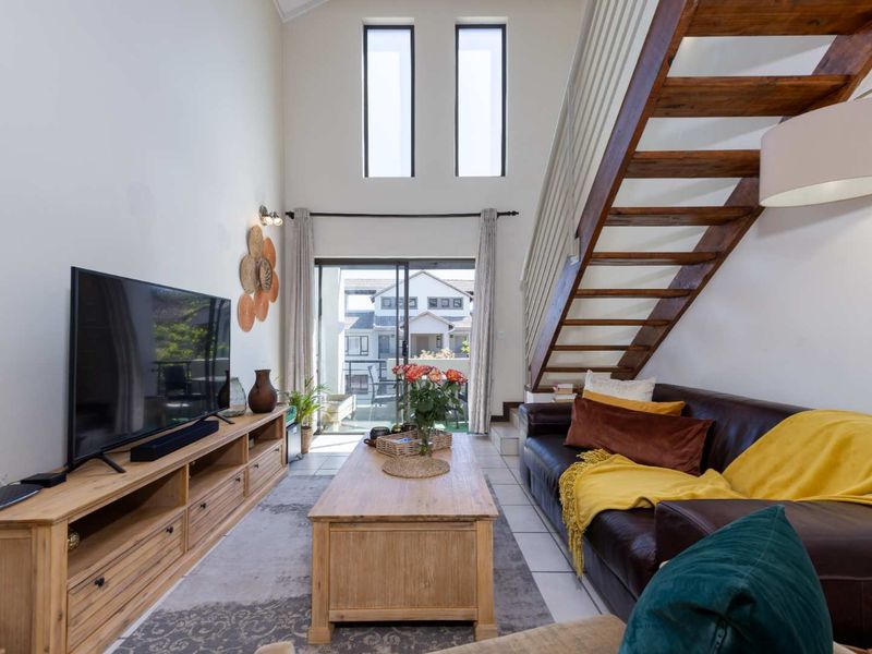 Bright double-volume living room featuring a timber staircase, tiled flooring, and large sliding glass doors leading to an outdoor area.