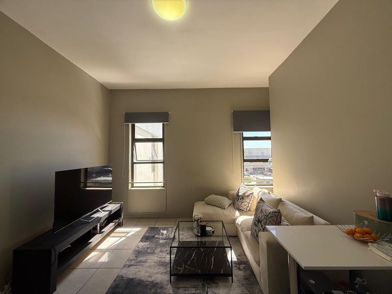 A well-lit living room featuring neutral wall tones, tiled flooring, and two windows with roller blinds, providing a comfortable and functional living space.