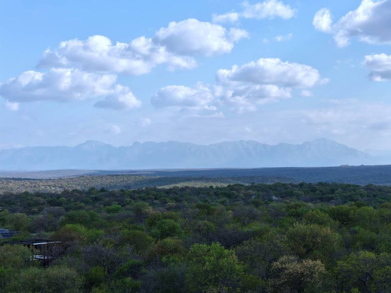 A scenic elevated view overlooking a vast expanse of natural bushveld and distant mountain ranges under a clear blue sky.