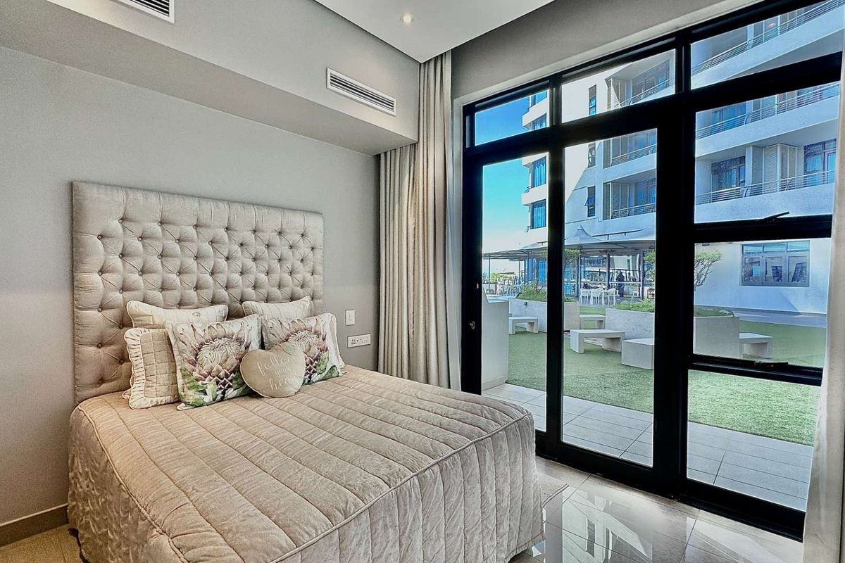 Modern bedroom featuring floor-to-ceiling glass sliding doors that open onto a private patio area, complemented by recessed ceiling lighting and ducted air conditioning.
