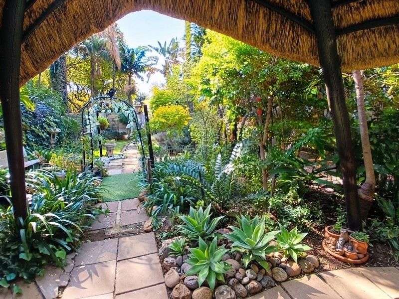 A lush, well-maintained garden featuring a stone pathway, metal archway, and mature landscaping, viewed from beneath a traditional thatched outdoor structure.