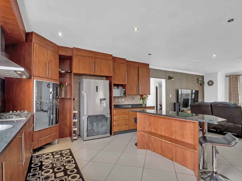 Open-plan kitchen and living area featuring warm-toned wooden cabinetry, granite countertops, built-in appliances, and neutral tiled flooring throughout.