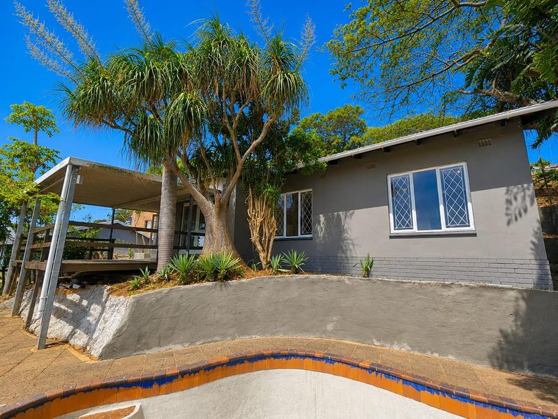Rear exterior view featuring a swimming pool, raised wooden deck, covered patio area, and mature landscaping against a grey painted brick facade.