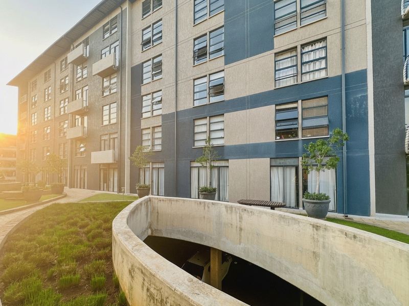Exterior view of a modern multi-story apartment building featuring balconies, a landscaped garden area, and a concrete ramp leading to underground parking.
