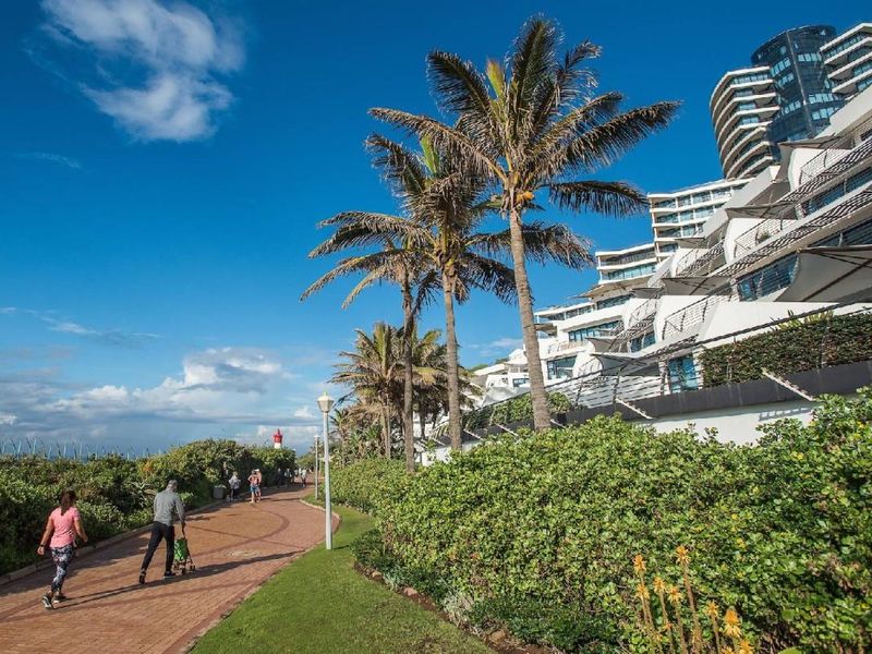 Exterior view of the apartment complex featuring tiered balconies, landscaped gardens, and a paved coastal walkway.