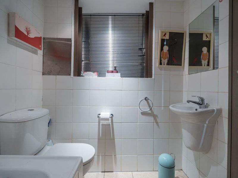 A functional bathroom featuring white ceramic wall tiles, a pedestal sink with chrome faucet, a standard toilet, and a window with venetian blinds.