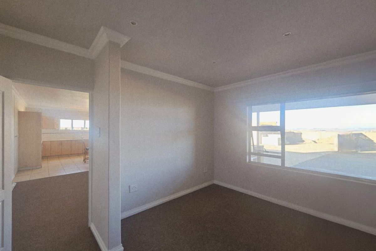 A spacious, empty living area featuring neutral carpeting, crown molding, recessed lighting, and a large window with views of the surrounding industrial exterior.