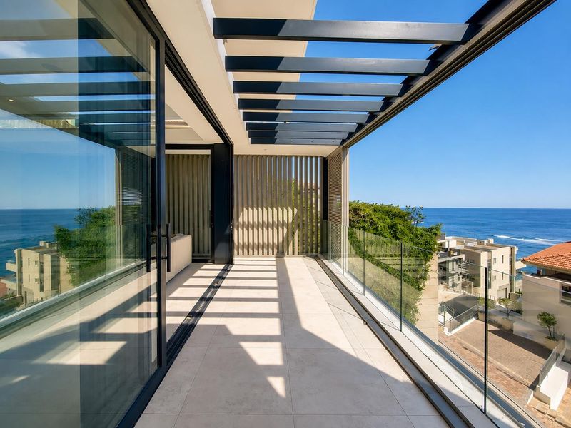 A modern, spacious balcony featuring a sleek glass balustrade, overhead pergola, and expansive sliding doors with stunning ocean views.