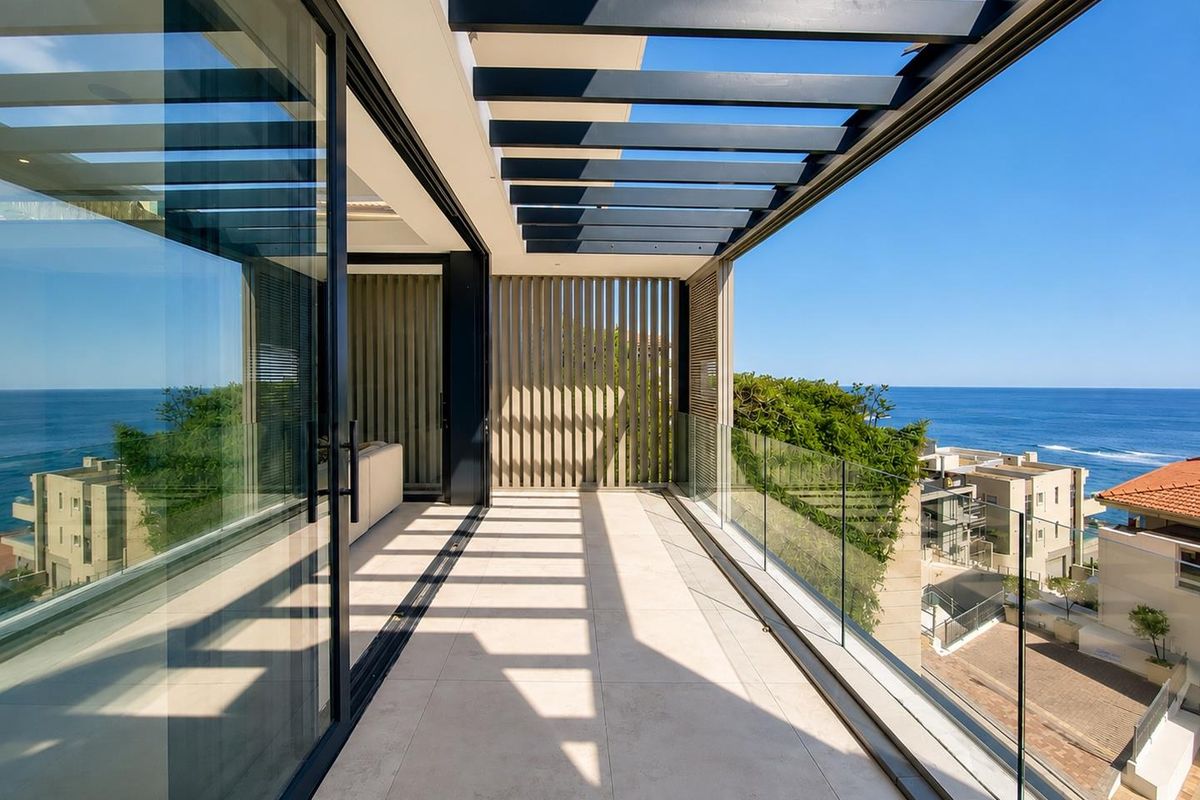 A modern, spacious balcony featuring a sleek glass balustrade, overhead pergola, and expansive sliding doors with stunning ocean views.