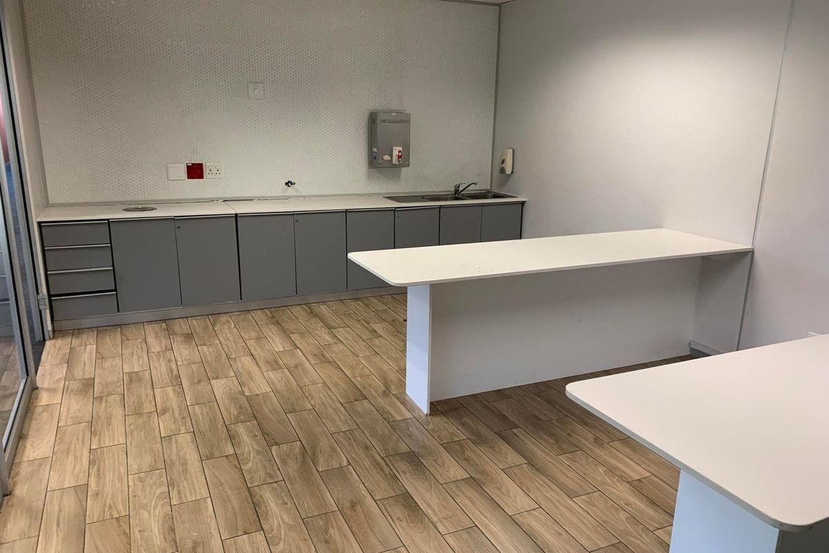 A modern office kitchenette featuring grey cabinetry, a white countertop, a stainless steel sink, and wood-look tile flooring.