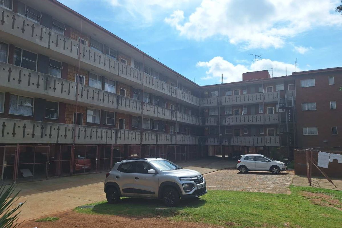 Exterior view of a multi-story apartment complex featuring brick facade, open-air walkways, and ground-level parking area.