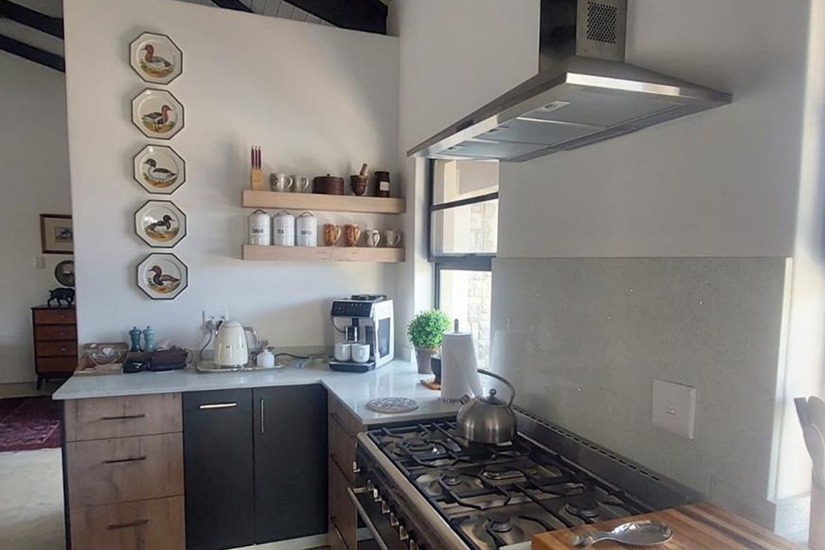 Modern kitchen featuring a stainless steel gas range, extractor fan, stone countertops, and floating timber shelves under an exposed beam vaulted ceiling.
