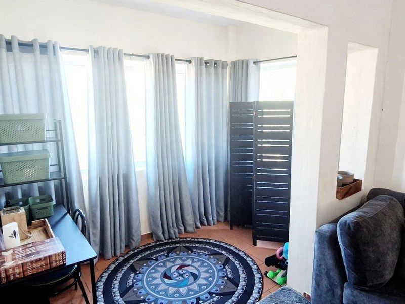 A bright living area featuring tiled flooring and large windows, providing ample natural light and a versatile space for relaxation or a home office setup.